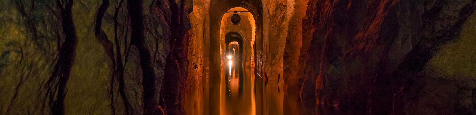 Fotografia przedstawia oświetlony chodnik w sztolni, na dnie którego znajduje się woda.