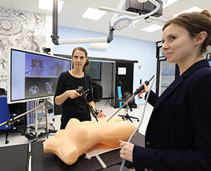 The photo shows two women performing a simulated procedure on a phantom in a modern operating theater.