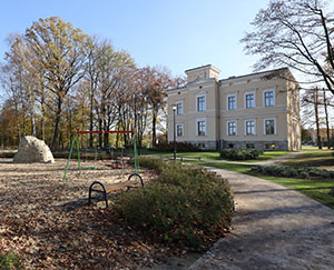 On the left side of the photo there is a section of the playground on the sandy ground with swings and a slide. On the right, there is a castle, an elegant building that has undergone extensive renovations.