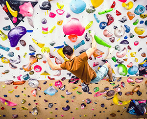 The photo shows a climbing wall with lots of colorful handholds and a young man in a brown T-shirt climbing.