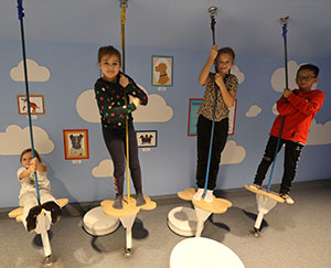The photo shows four children – three girls and a boy – swinging on balances suspended with a rope and attached to the ground.