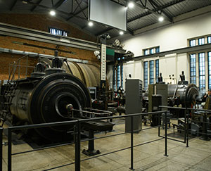 In the photo there is a hall where two steam engines have been installed, with a large rope wheel behind them. The control panels are also visible.
