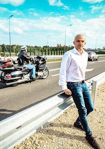 Fotografia przedstawia Piotra Holdenmajera, mieszkańca Rybnika. Łysy mężczyzna w białej koszuli i dżinsach siedzi na barierce energochłonnej przy drodze. Za nim znajduje się ulica, po której porusza się między innymi motocyklista. Autorką zdjęcia jest Lucyna Nenow.