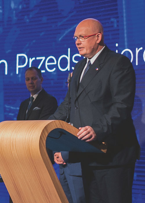 Michał Gramatyka, wicemarszałek województwa śląskiego, przemawia, stojąc za pulpitem. Z lewej strony widać stolik konferencyjny i stojącego kolejnego prelegenta.