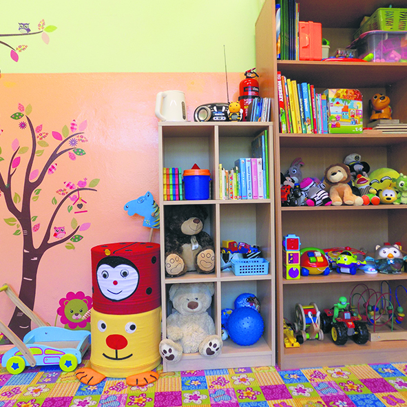Na kolorowej ścianie umieszczony jest regał z licznymi półkami, na których znajdują się zabawki dla dzieci: pluszaki, lalki, klocki, książki i gry.