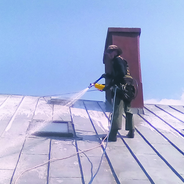 Mężczyzna ubrany w roboczy ochronny uniform stoi na dachu i maluje go metodą natryskową.