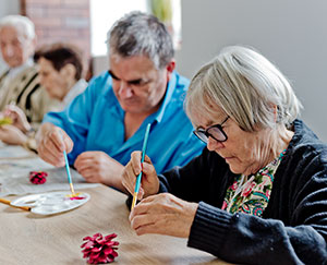 The photograph shows three senior citizens creating ornaments during an art class.