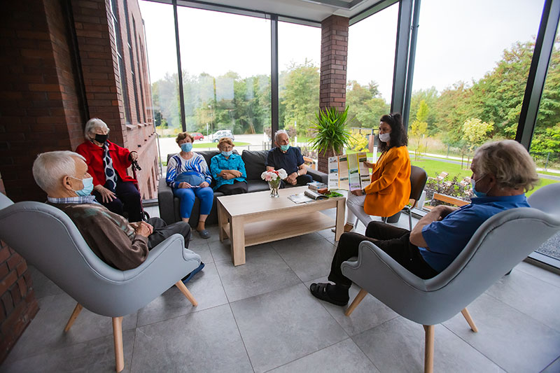 Fotografia przedstawia sześcioro seniorów siedzących na kanapach, wokół stolika, w trakcie zajęć. Prowadzi je kobieta w żółtej marynarce. Ściany są przeszklone, widoczny jest ogród i fragment parkingu.