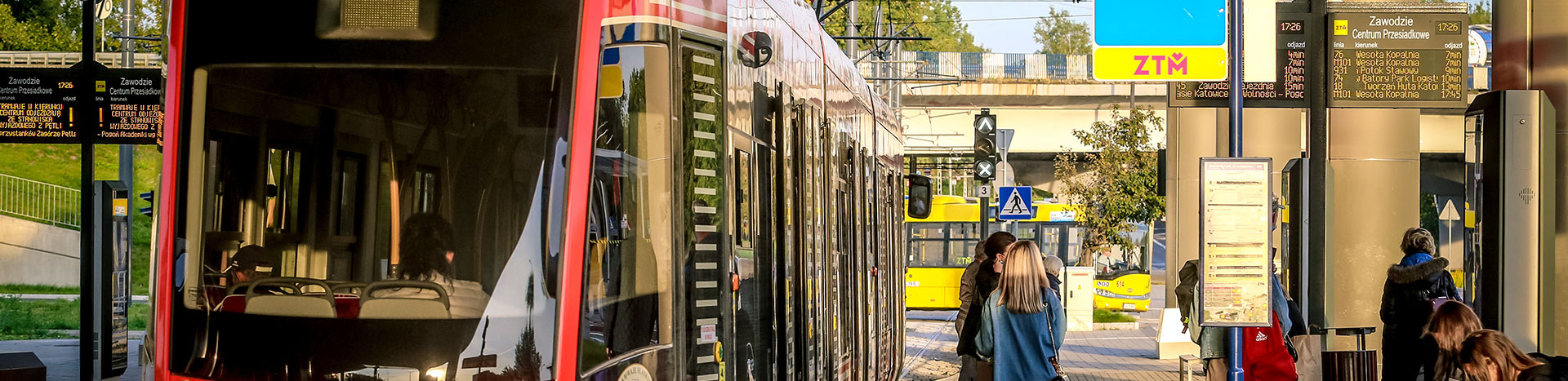 Fotografia przedstawia tramwaj  i ludzi na zadaszonym przystanku.