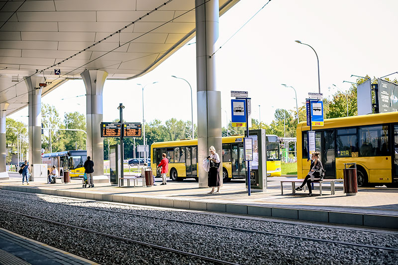 Fotografia przedstawia peron, przy którym stoją trzy autobusy. Na przystanku stoi kilku oczekujących pasażerów.