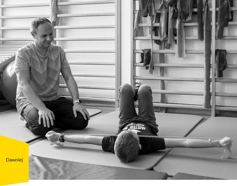 Fotografia przedstawia chłopca leżącego na materacu na sali gimnastycznej, który ćwiczy wraz z fizjoterapeutą, klęczącym obok.
