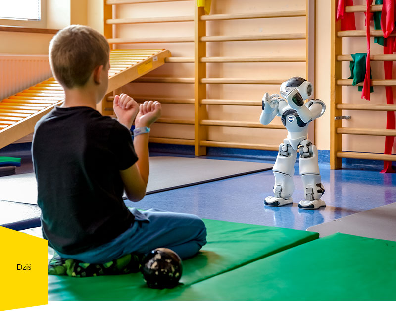 Fotografia przedstawia chłopca klęczącego na materacu na sali gimnastycznej, który wykonuje ćwiczenia prezentowane przez robota.