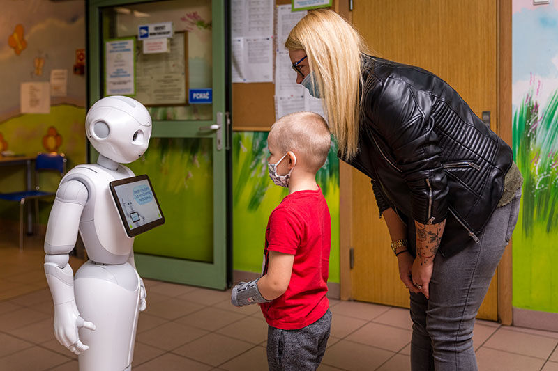 Fotografia przedstawia małego chłopca, który stoi przed robotem i wpatruje się w niego. Nad chłopcem pochyla się mama.
