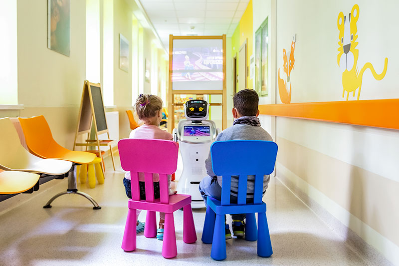 Fotografia przedstawia dwójkę dzieci siedzących na krzesłach, które oglądają wideo wyświetlane na ekranie robota.