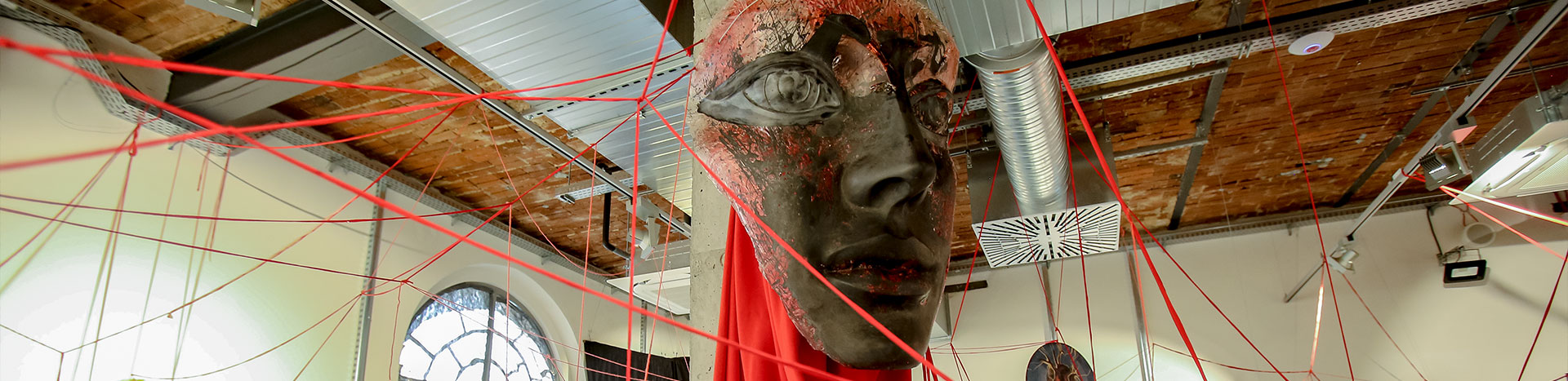 Fotografia przedstawia element ekspozycji. To maska, zawieszona wokół czerwonych sznurków.
