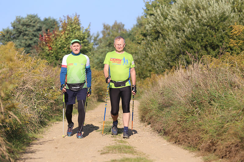 Fotografia przedstawia dwóch mężczyzn, ubranych na sportowo, idących polną ścieżką z kijkami do nordic walking.