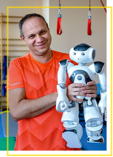 Fotografia przedstawia Łukasza Mierzwę, Koordynatora Zespołu Fizjoterapeutów na Oddziale Rehabilitacji Centrum Pediatrii im. Jana Pawła II w Sosnowcu. Mężczyzna, stojąc w sali gimnastycznej,  trzyma w rękach robota.