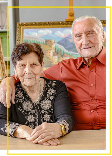 Fotografia przedstawia Romana Chromego, uczestnika projektu. Starszy mężczyzna obejmuje starszą kobietę. Za nimi znajdują się sztalugi z obrazami.