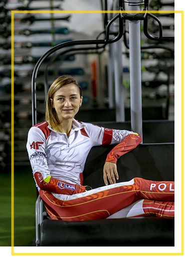 Fotografia przedstawia Monikę Hojnisz-Staręgę, polską biathlonistkę, medalistkę mistrzostw świata i Europy. Narciarka w biało-czerwonym kombinezonie, siedzi na ławce stylizowanej na krzesełko wyciągu.