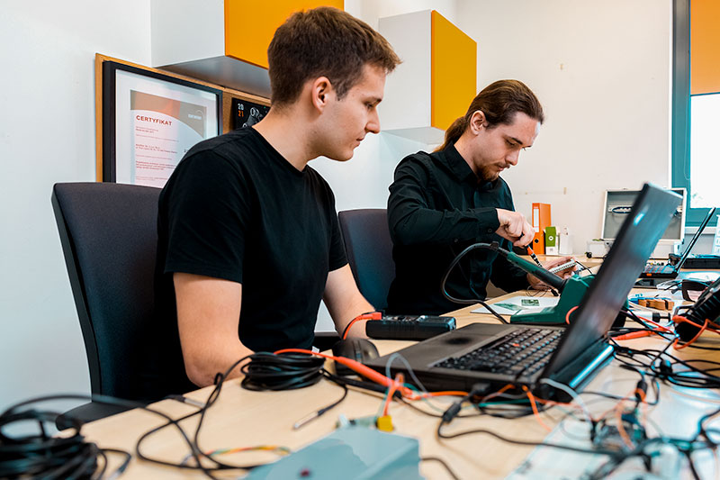 Fotografia przedstawia dwóch mężczyzn, którzy programują i składają specjalne czujniki. Na stole widoczne są komputery, kable i narzędzia.