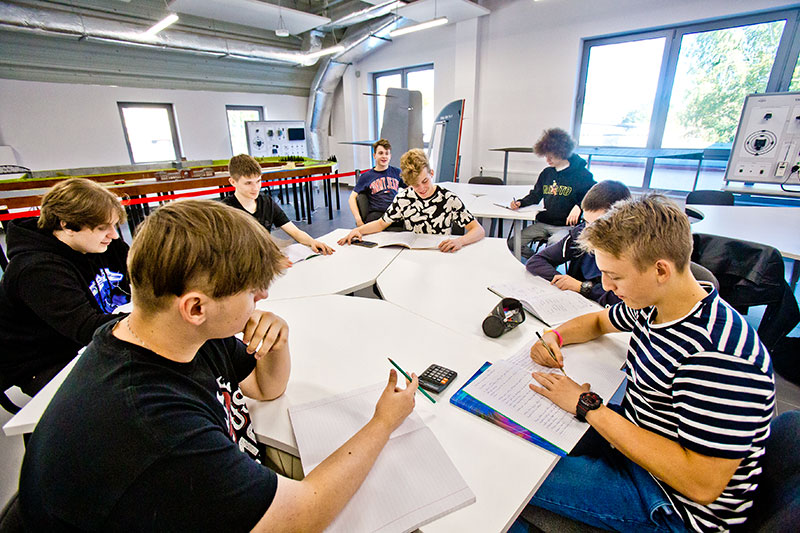 Fotografia przedstawia kilku uczniów siedzących przy okrągłym stole, notujących coś w zeszytach.