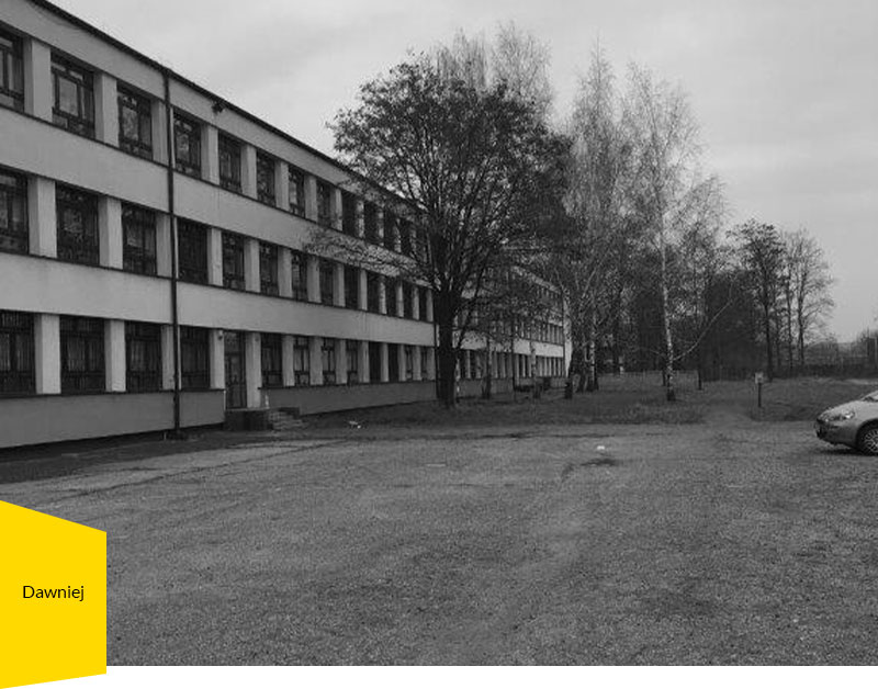 Fotografia przedstawia budynek szkolny w kształcie długiego prostopadłościanu oraz żwirowy parking.