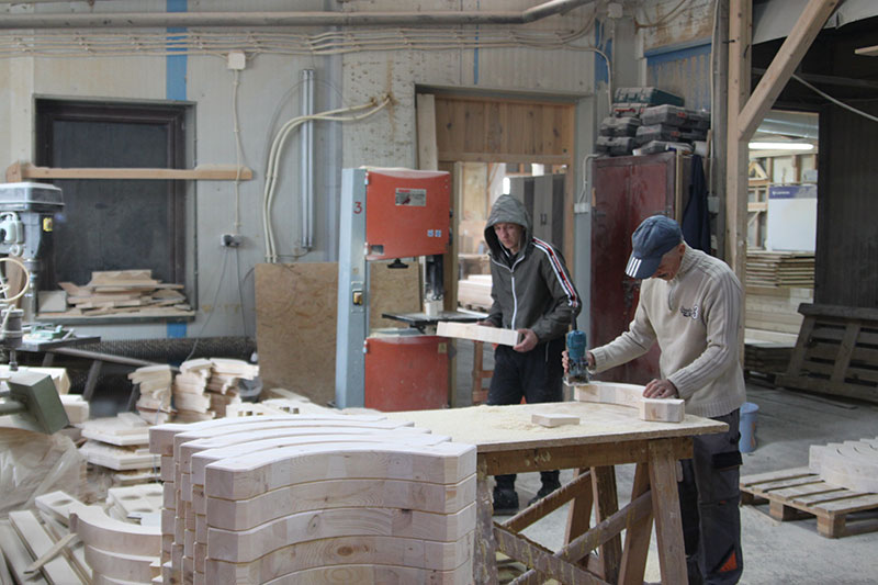 Fotografia przedstawia dwóch pracowników zakładu stolarskiego, przy pracy z drewnem.