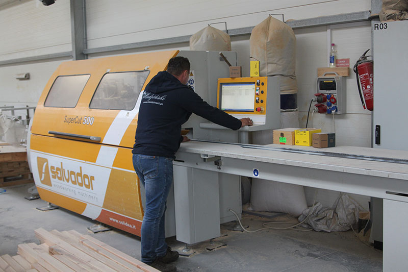 Fotografia przedstawia dużą maszynę do obróbki drewna obsługiwaną przez pracownika zakładu stolarskiego.