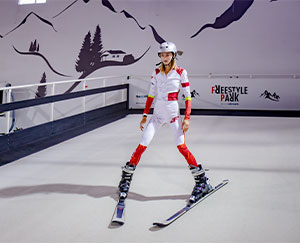 The photograph shows a young woman in a red and white jumpsuit skiing down an indoor slope.