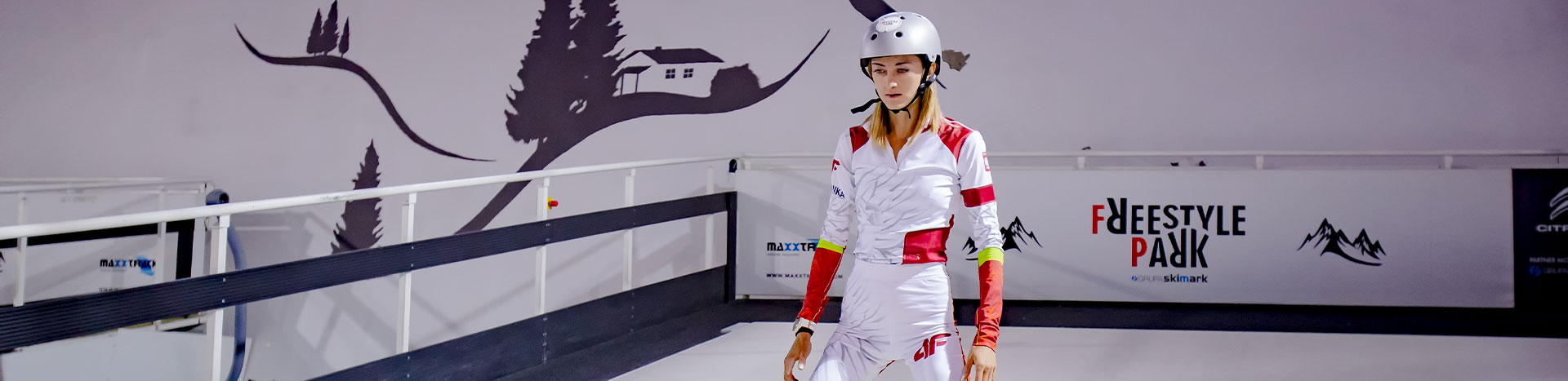 Fotografia przedstawia młodą kobietę w biało-czerwonym kombinezonie, która zjeżdża na nartach, na halowym stoku.