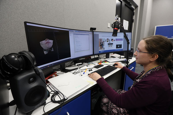 Na zdjęciu widzimy kobietę obsługującą komputer z trzema monitorami. Wyświetlone są na nich różne dane i informacje, między innymi fragment ludzkiej twarzy.