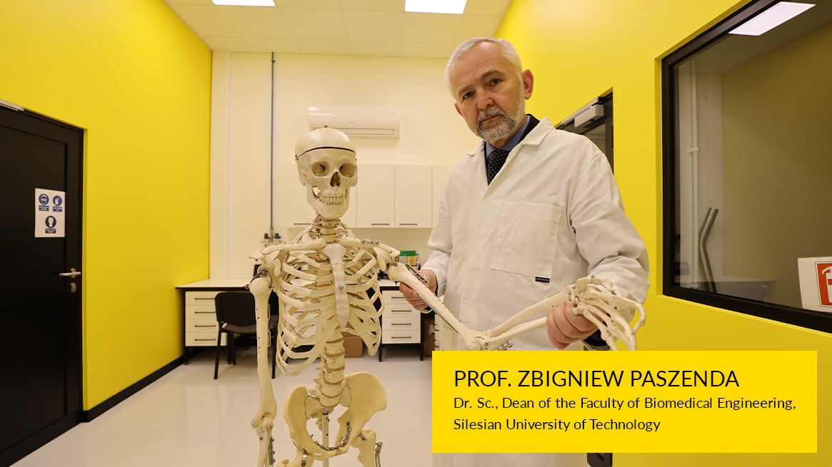The photograph shows Zbigniew Paszenda. A middle-aged man with short-cropped gray hair and a beard is wearing a doctor's apron. He is holding the hand of the skeleton model.