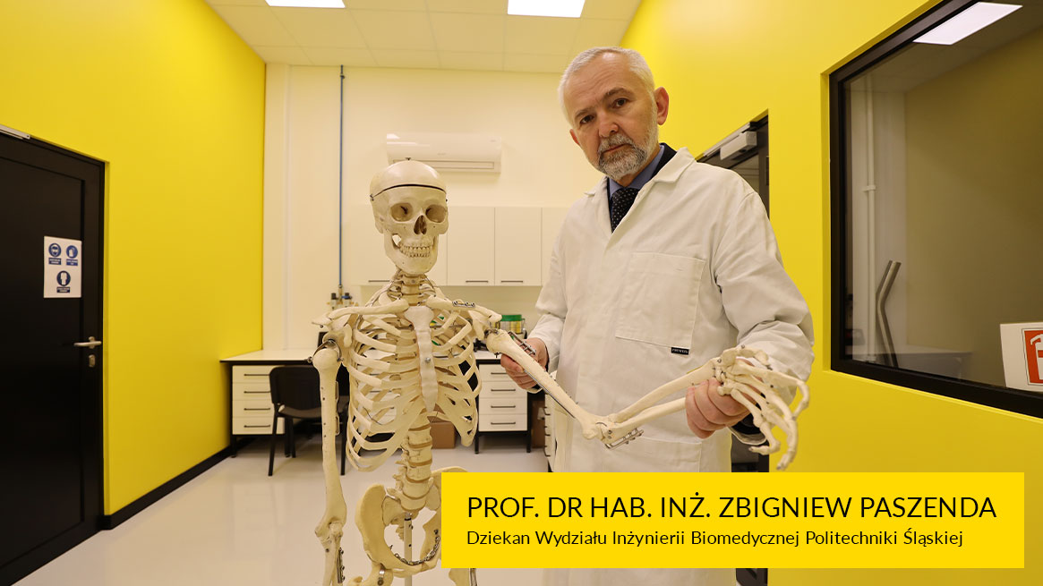 Fotografia przedstawia Zbigniewa Paszendę. Mężczyzna w średnim wieku o krótko przystrzyżonych, siwych włosach i zaroście, jest w lekarskim fartuchu. Trzyma rękę modelu szkieletu.