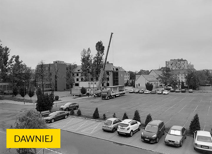Zdjęcie przedstawia plac budowy, przed rozpoczęciem robót. Widoczne są zaparkowane wokoło samochody oraz dźwig stojący z lewej strony betonowego placu.
