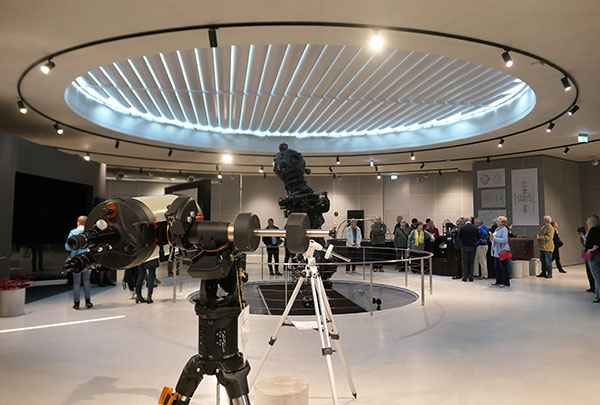 The photograph shows a group of people visiting a planetarium. There are devices on display which look like telescopes.