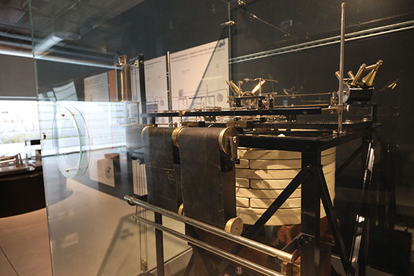 Fotografia ukazuje maszynę na ekspozycji. Widzimy między innymi rolki czy inne skomplikowane elementy. Znajduje się w szklanej gablocie.