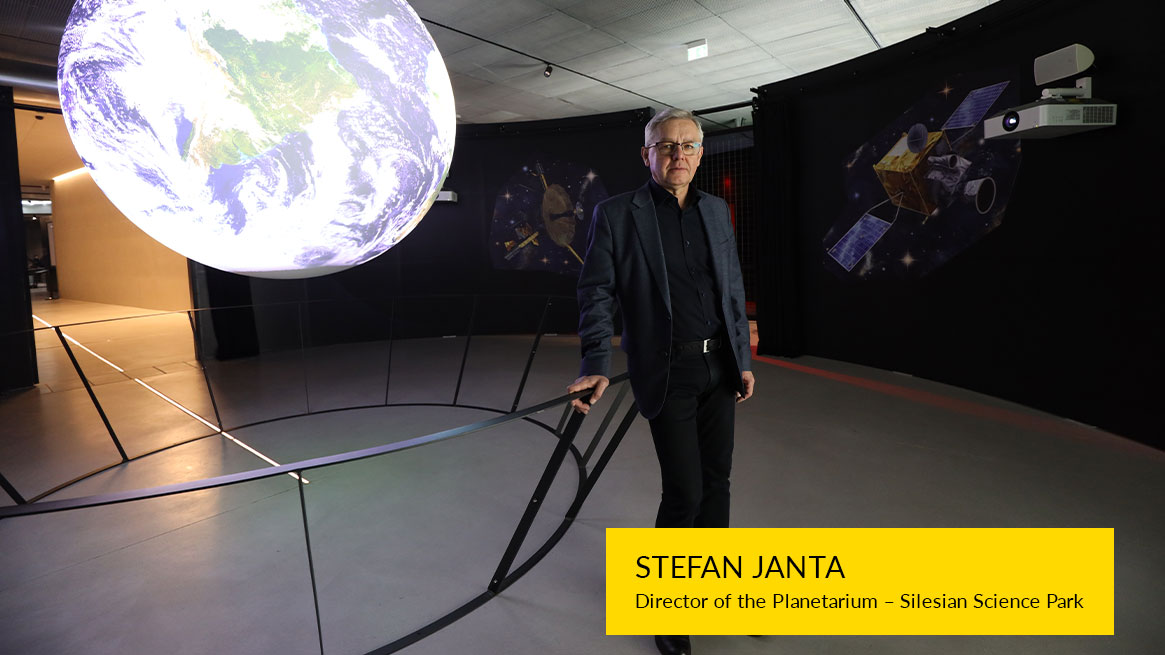 The photo shows a man with gray hair, glasses, wearing a dark shirt and a jacket. He is standing, leaning against a railing, with a model of the Earth in the background.