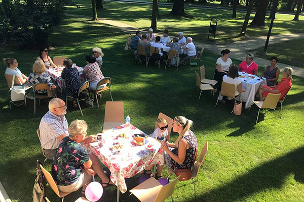 Fotografia przedstawia uczestników rodzinnego pikniku. Przy czterech stołach ustawionych na trawie, siedzą kobiety i mężczyźni w różnym wieku.