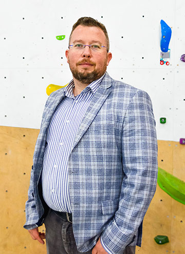 The photograph shows a man wearing glasses and an unbuttoned light-colored jacket. A section of the climbing wall can be seen in the background.