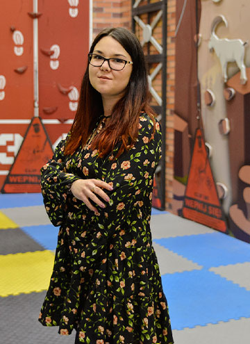 The photo shows a smiling long-haired woman with glasses who is wearing a dress. She has her arms folded. Climbing walls for children are visible in the background.
