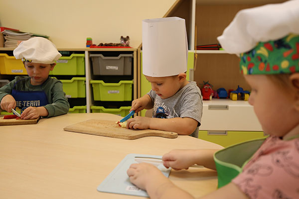 Fotografia przedstawia zajęcia kulinarne w przedszkolu. Widoczna jest trójka dzieci z czapkami kucharskimi. Wszystkie w skupieniu kroją owoce plastikowymi nożykami na drewnianych deskach.
