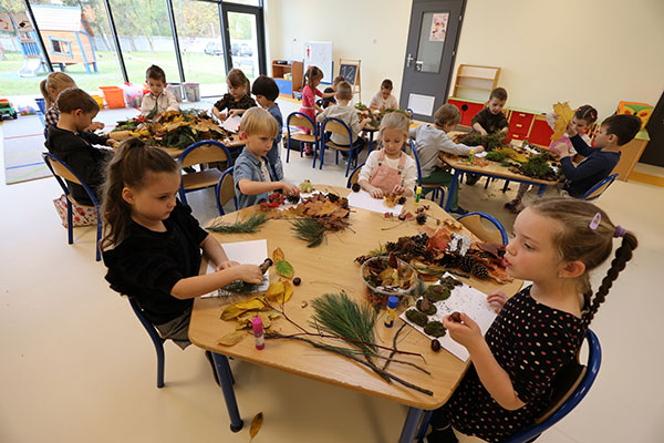 Zdjęcie przedstawia zajęcia plastyczne w przedszkolu. Dzieci siedzą przy kilku stolikach i wykonują pracę między innymi za pomocą liści.