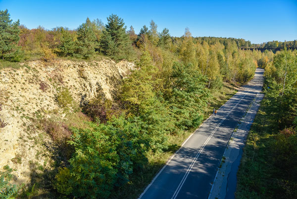 Fotografia wykonana jest z mostu, który biegnie nad wąwozem z drogą. Po lewej stronie widać skalną ścianę oraz mnóstwo drzew. Ulicą spacerują dwie osoby.