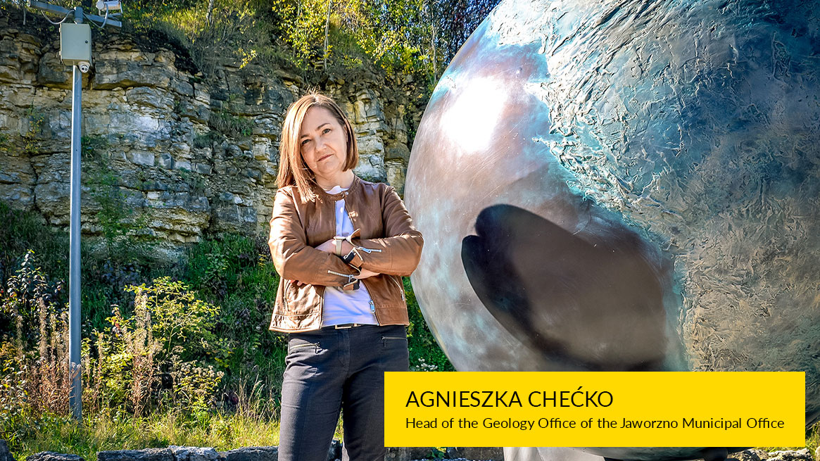 In the photo there is a woman in a brown leather jacket with her hands folded. She is standing next to a globe sculpture, with a rock wall behind her.