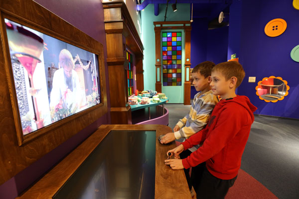 Na zdjęciu widzimy dwóch chłopców biorących udział w interaktywnej bajce. Znajdują się w kolorowym pomieszczeniu. Stoją przed monitorem, a postaciami sterują za pomocą specjalnych dźwigni.