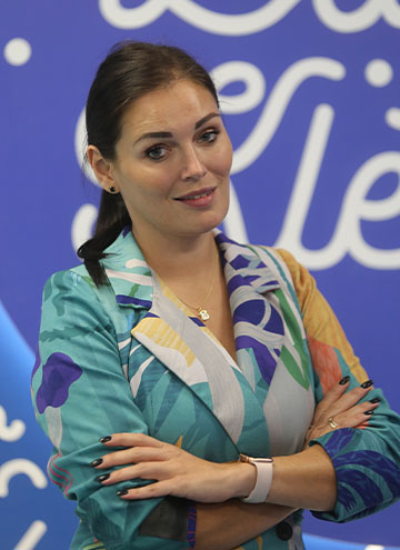 The photograph shows a woman with her black hair pinned up and her hands folded. She is wearing a jacket with a floral pattern.