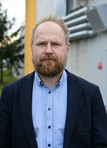 The photo shows a man with a thick beard wearing a dark jacket and shirt. There is a technical building behind him.