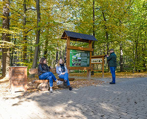 Na zdjęciu widoczne są dwie rozmawiające kobiety, siedzące na ławce w parku. Obok znajduje się tablica informacyjna, której przygląda się mężczyzna w kaszkiecie i kurtce.
