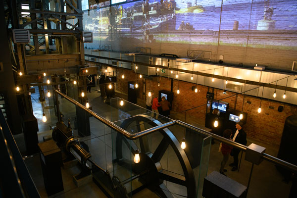 Na zdjęciu wykonanym z góry, widzimy osoby zwiedzające wystawę. Widocznych jest kilka interaktywnych pulpitów, szyb windy kopalnianej, rury czy koło szybowe.