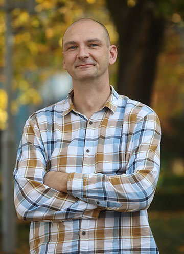 The photograph shows a smiling man with short cropped hair, wearing a plaid shirt, with his arms folded. 